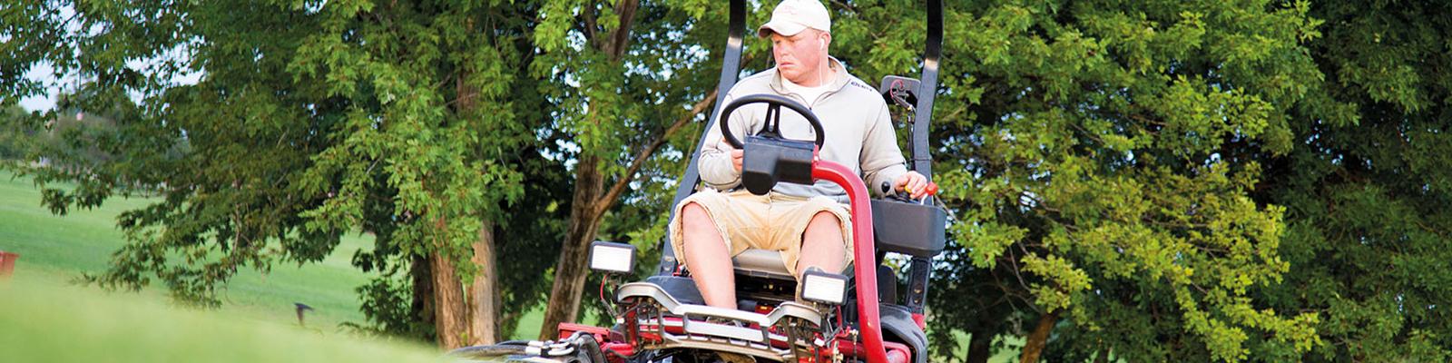 Man on large riding lawnmower, trimming grass on a golf course.